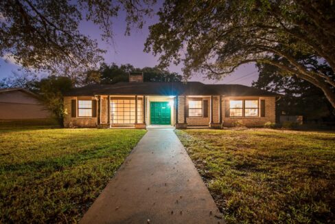 Front view of a brick house with a green door and large windows, illuminated at dusk.