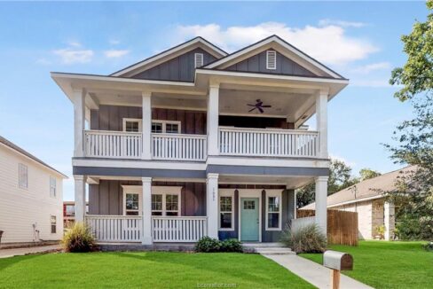 Two-story house with a large front porch and white railings in a suburban neighborhood.