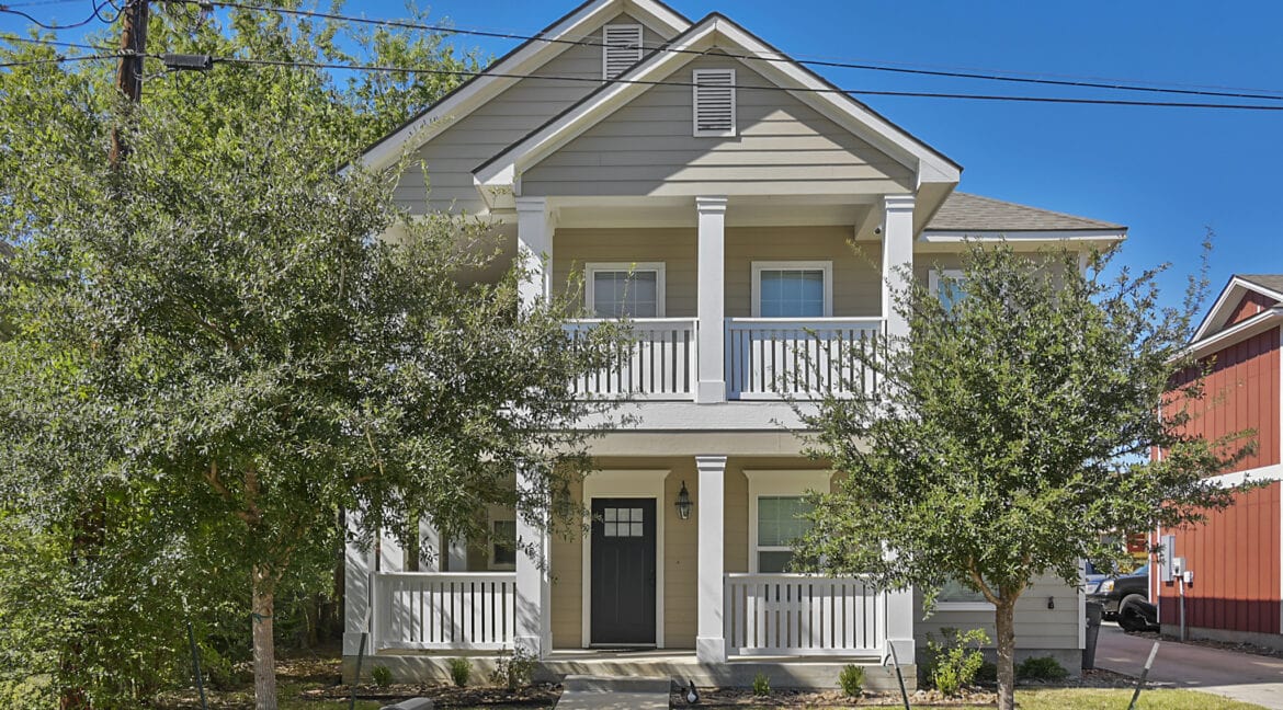 Front view of a two-story house with a porch and white railing, surrounded by trees in Aggieland.