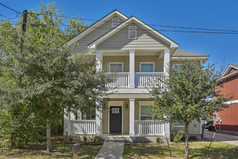 Front view of a two-story house with a porch and white railing, surrounded by trees in Aggieland.