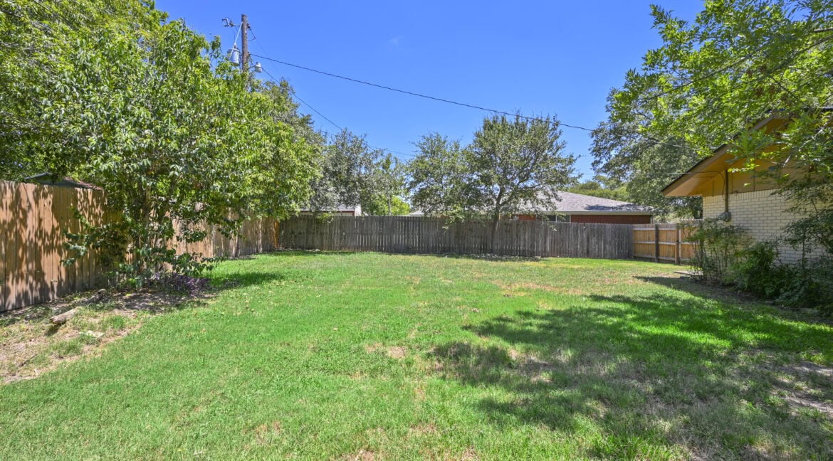 Spacious backyard with green grass, trees, and a wooden fence in a residential area.