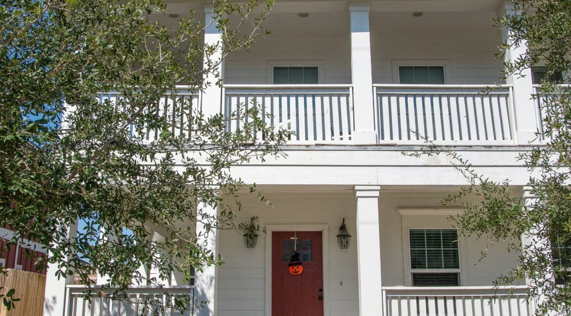 Three-story white house with a red front door and a small front yard under a clear blue sky.