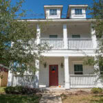 Three-story white house with a red front door and a small front yard under a clear blue sky.